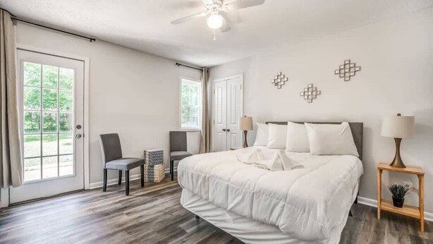 A bedroom with a large bed, a ceiling fan, two bedside tables with lamps, a chair, and a window with curtains. - Judy Lynn Collective (Memphis)