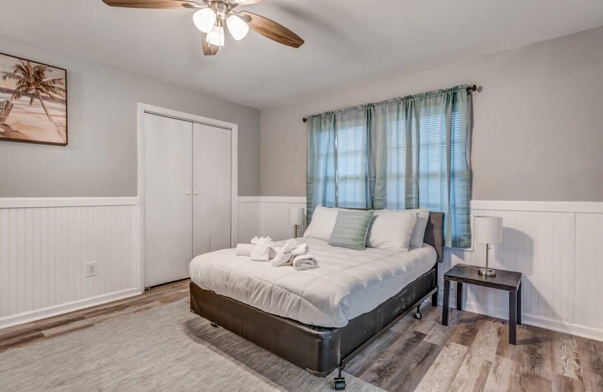 A bedroom with a bed, a ceiling fan, a window with curtains, a bedside table with a lamp, and a door.