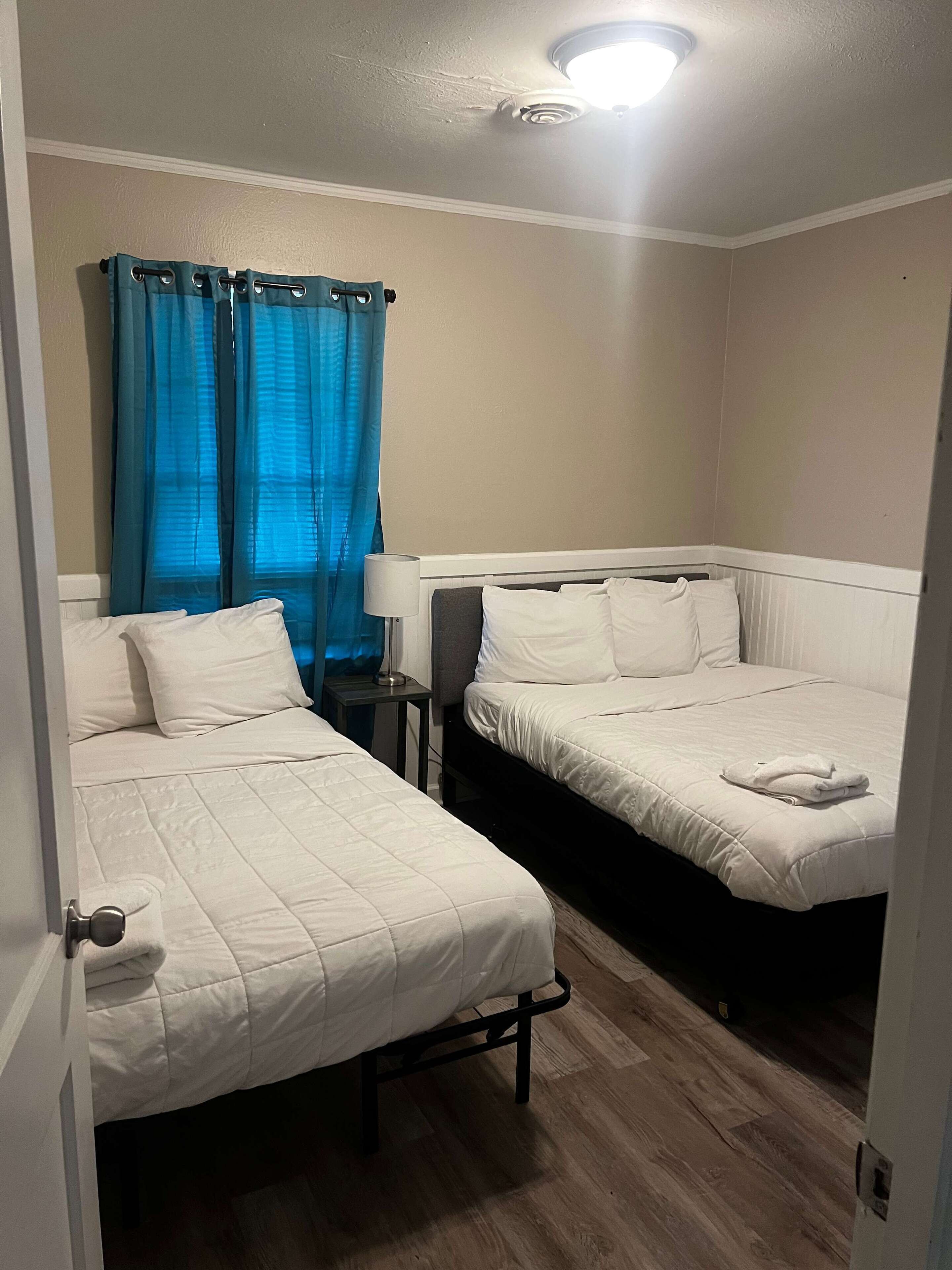 Een kamer met twee bedden, een nachtkastje, een lamp en een raam met blauwe gordijnen.