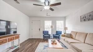 Una sala de estar con ventilador de techo, televisor, dos sillones azules, un sofá beige y una mesa de centro con bandeja.