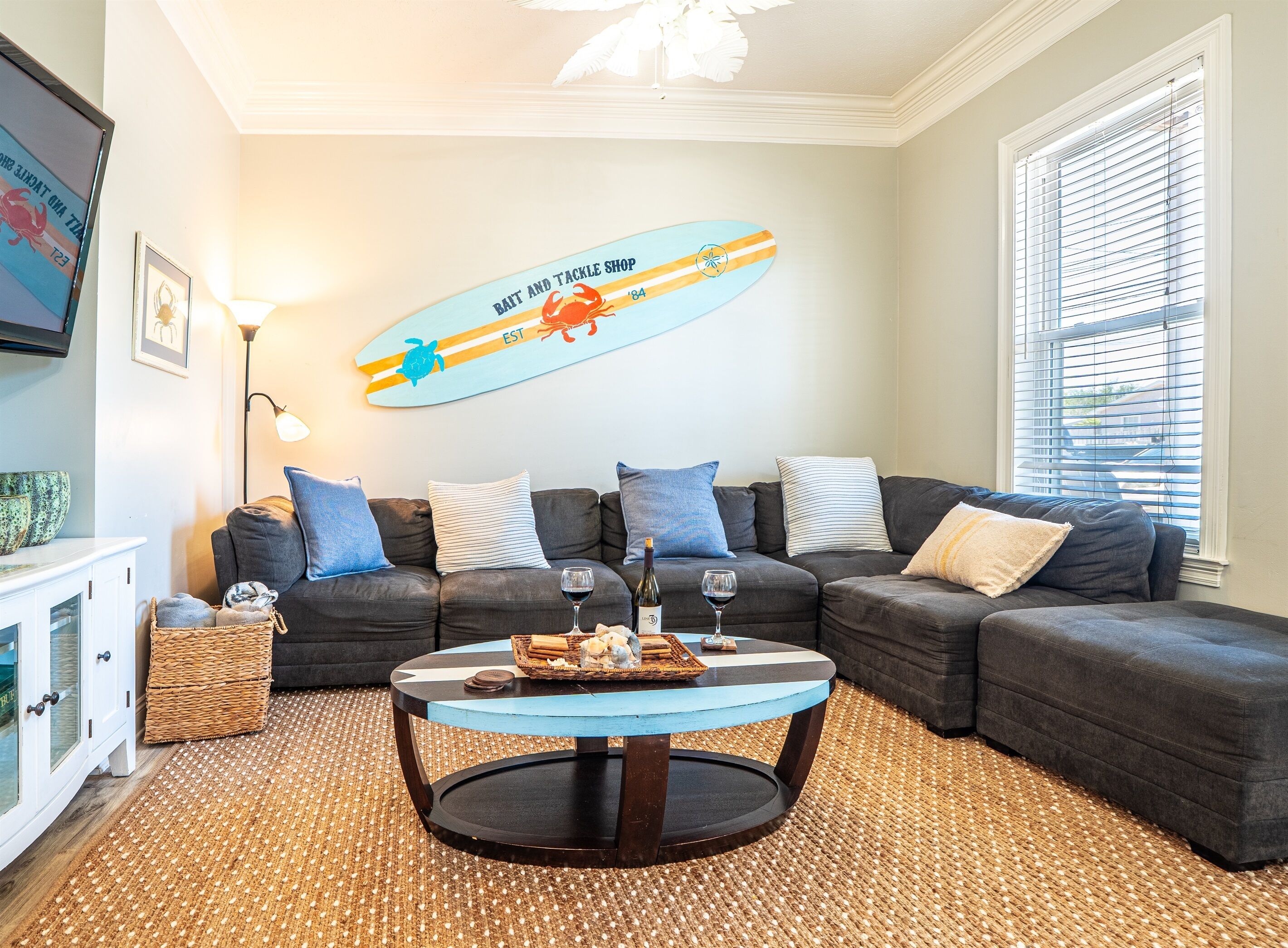 Een woonkamer met een donkergrijze hoekbank, een ronde salontafel met een geweven onderzetter, en een surfboard decoratie aan de muur.