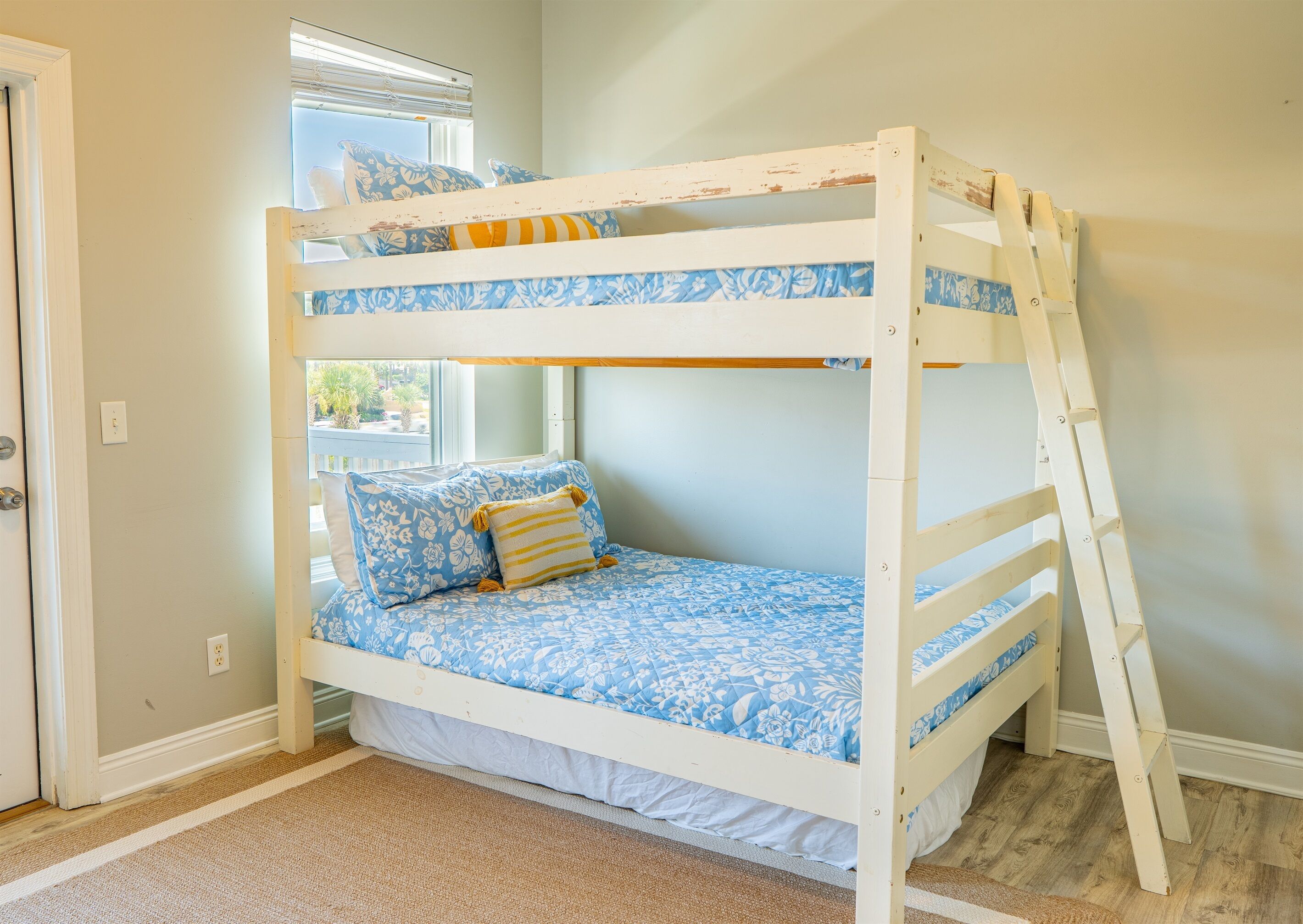 A bunk bed with floral bedding, a window with blinds, and a wooden ladder.
