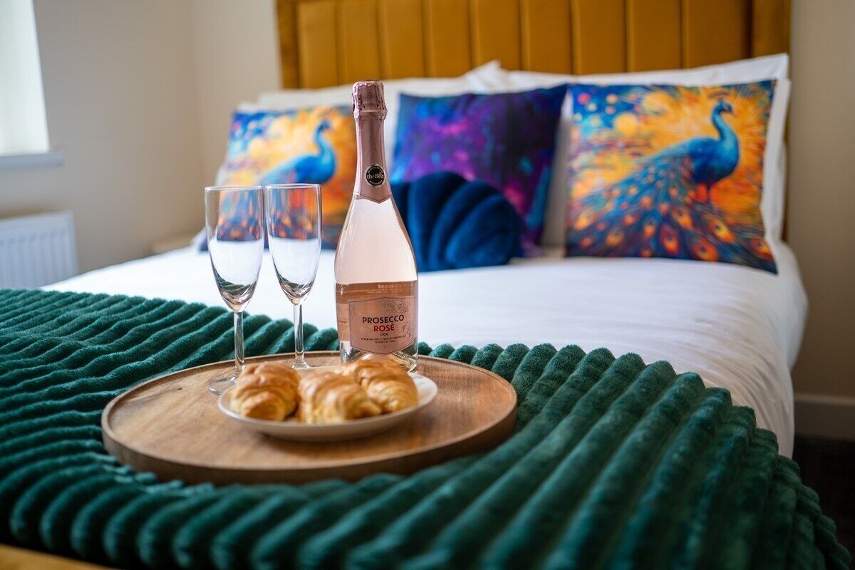 Een bed met een groene deken, een houten dienblad met croissants en champagne, en twee glazen.