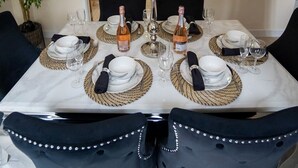 A dining area with a table set for four, featuring white plates, clear glasses, and woven placemats. There are two bottles of wine, a candelabra with candles, and a television mounted on the wall displaying a fireplace. - Luxury 5BR Home | Hot Tub | EV-Friendly Stay (West Bromwich)