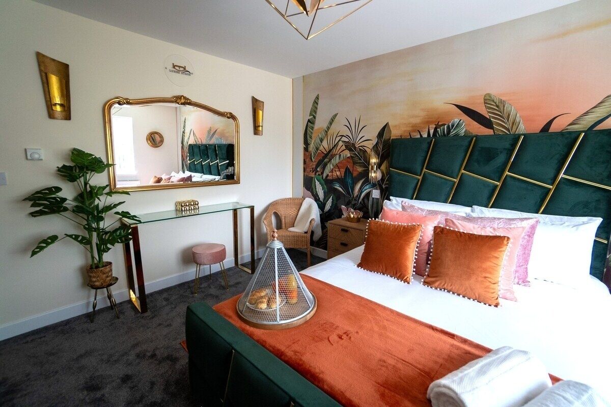 Ein Hotelzimmer mit einem großen Bett, einem grünen, gepolsterten Kopfteil, einem Spiegel, einem Schreibtisch und einer Zimmerpflanze.