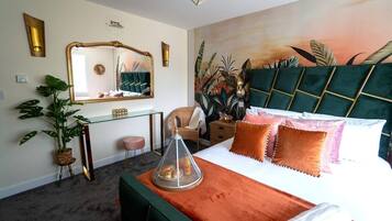 Ein Hotelzimmer mit einem großen Bett, einem grünen, gepolsterten Kopfteil, einem Spiegel, einem Schreibtisch und einer Zimmerpflanze.