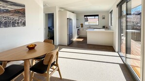 A modern kitchen with a central island, a dining area with a table and chairs, and a large window providing a view of the outdoors. - Roto Cottage - Lake Tekapo (Lake Tekapo)