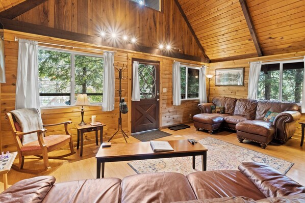A cozy cabin interior with wooden walls, a leather sofa, a rocking chair, and a dining table.