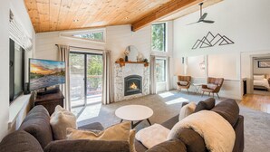 A cozy living room with a fireplace, large windows, and a ceiling fan.