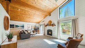 A spacious living room with a stone fireplace, wooden ceiling, and large windows offering a view of trees.