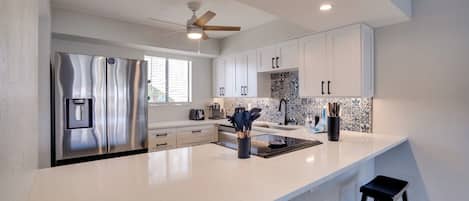 A modern kitchen with a stainless steel refrigerator, white cabinets, a black backsplash, and a ceiling fan.
