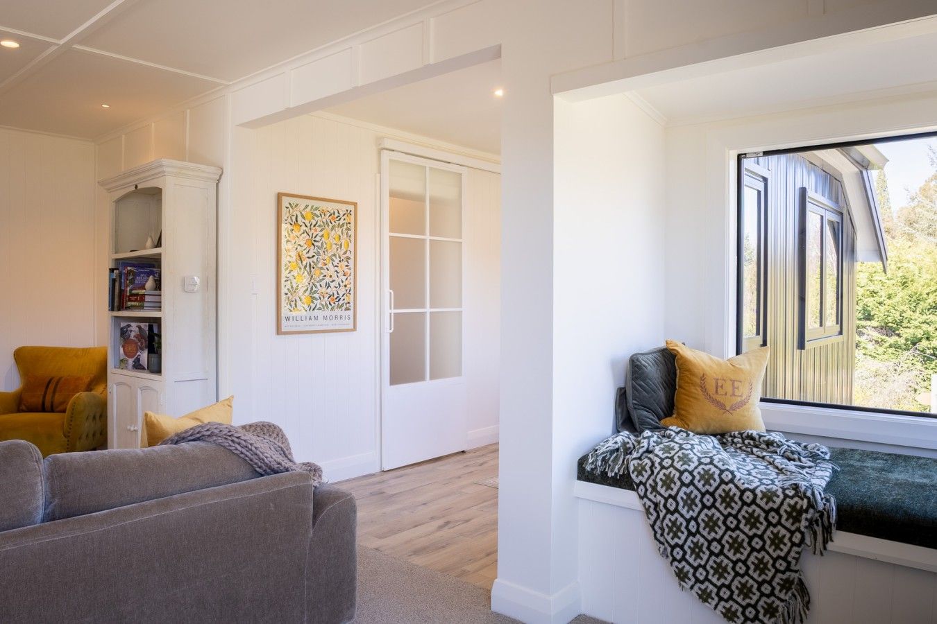 A room with a grey sofa, a yellow armchair, a bookshelf, a framed artwork, and a window with a patterned throw.