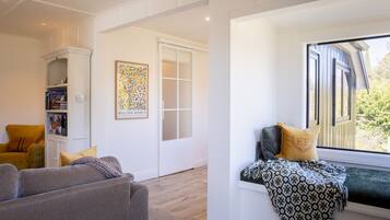 A room with a grey sofa, a yellow armchair, a bookshelf, a framed artwork, and a window with a patterned throw.