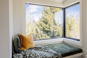 Ein Zimmer mit einem großen Fenster mit Blick auf einen Wald, eine gepolsterte Bank mit einem gemusterten Überwurf und ein Kissen.