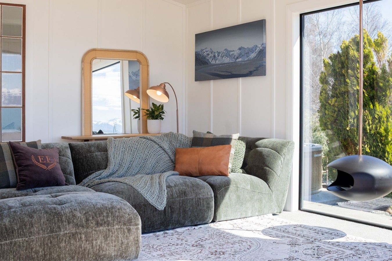 En moderne stue med en hjørnesofa, et stort spejl, et maleri og udsigt til træer gennem et stort vindue.