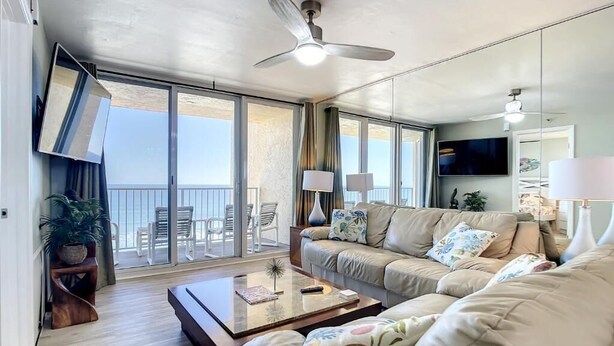 A modern living room with a sofa, coffee table, and a TV. There is a balcony with a view of the sea.