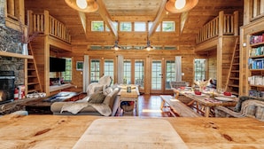 A spacious log cabin interior with a fireplace, wooden furniture, and a large dining table. - NEW! Views of Whitefish Lake, Wood Burning Fireplace, Cozy Cabin (Whitefish)