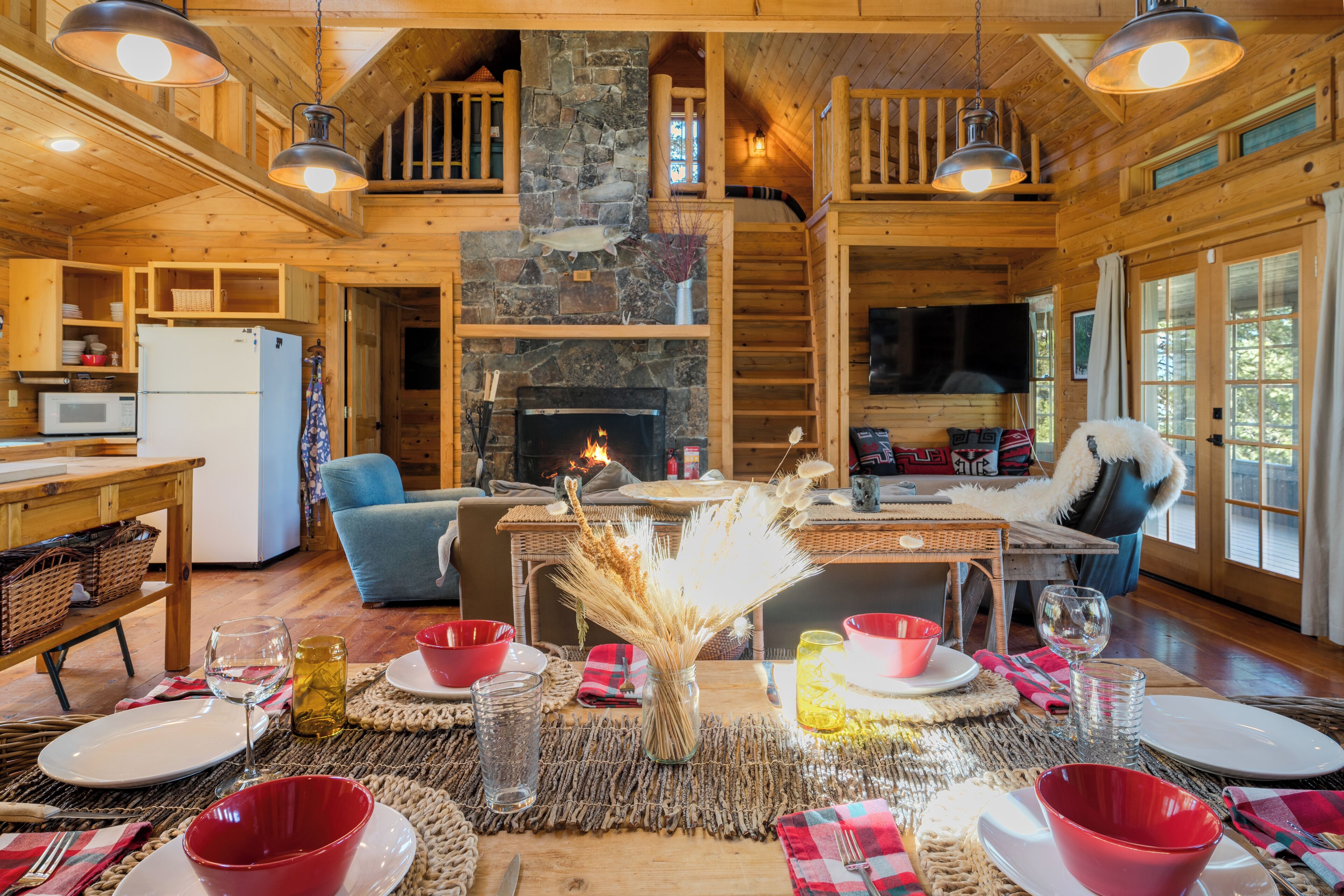 A cozy cabin interior with a stone fireplace, wooden dining table, and a loft bedroom.