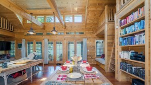 A wooden cabin interior with a dining area, bookshelves, and a large window. - NEW! Views of Whitefish Lake, Wood Burning Fireplace, Cozy Cabin (Whitefish)