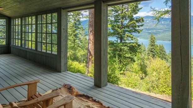 A wooden deck with Adirondack chairs and a fur rug, overlooking a forest and lake. - NEW! Views of Whitefish Lake, Wood Burning Fireplace, Cozy Cabin (Whitefish)
