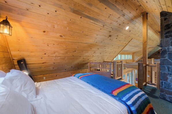 Acogedora habitación tipo cabaña de madera, con cama, manta colorida, chimenea de piedra y escalera de madera.