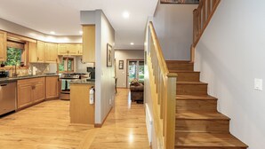 A spacious kitchen with wooden cabinets and a central island, a wooden staircase, and a view of the outdoors through a window. - Mountain Harbor Hideaway! Walking distance to Whitefish Lake (Whitefish)