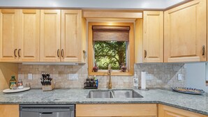A kitchen with wooden cabinets, a granite countertop, a stainless steel sink, and a window with a view of trees. - Mountain Harbor Hideaway! Walking distance to Whitefish Lake (Whitefish)