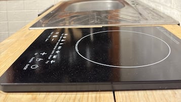 A kitchen with a black induction cooktop, a wooden countertop, and a shelf above containing glasses and kitchen utensils.
