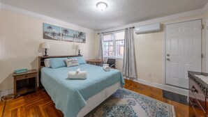 Ein Schlafzimmer mit Bett, Nachttisch, Lampe, Fenster mit Vorhängen, Tür und einer Wand mit zwei Bildern.