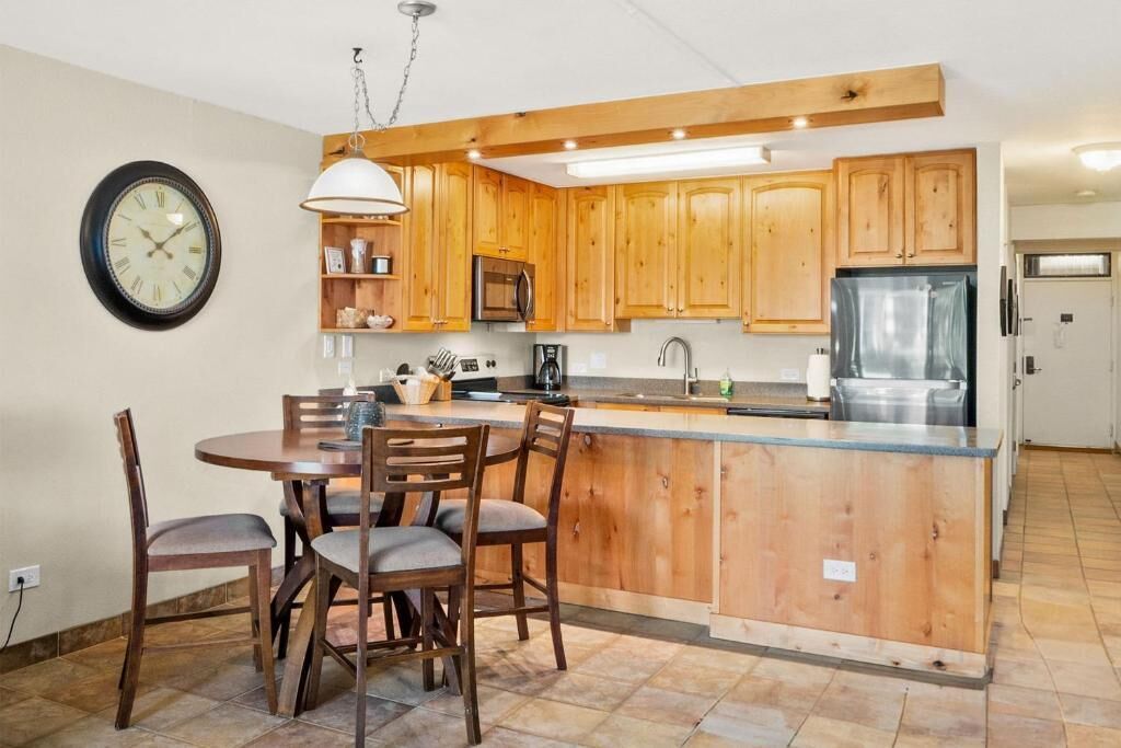 Une cuisine avec des meubles en bois, un îlot central, une table à manger avec des chaises, une horloge au mur et un luminaire suspendu.