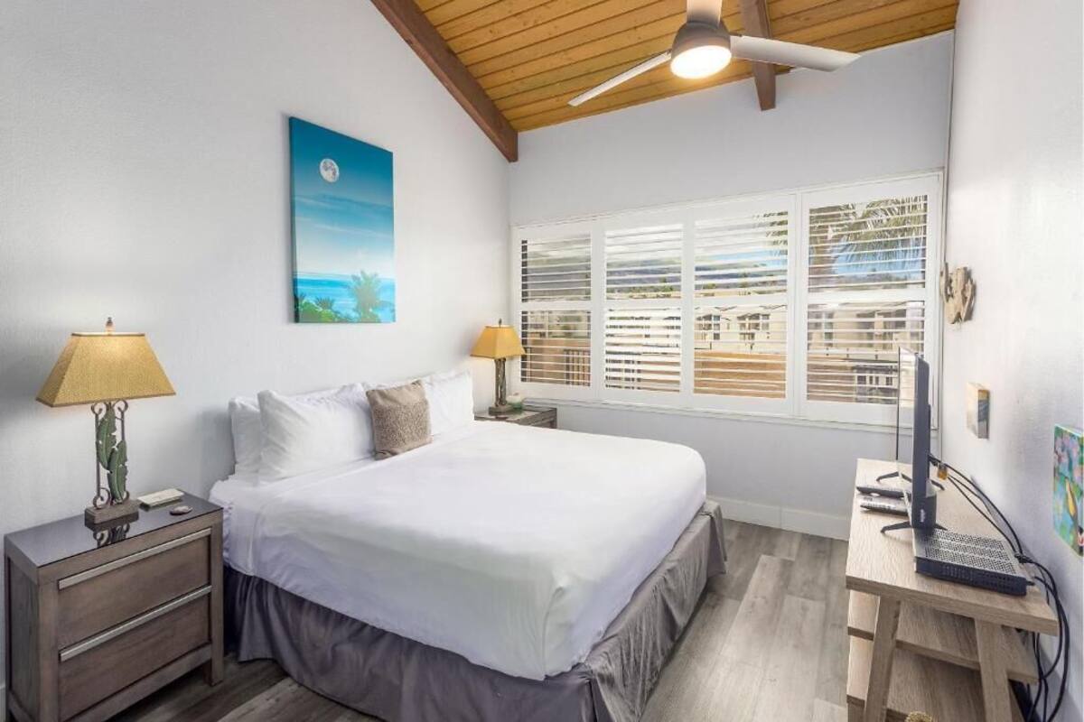A bedroom with a bed, two bedside tables with lamps, a desk with a computer, a ceiling fan, a window with blinds, and a painting on the wall.