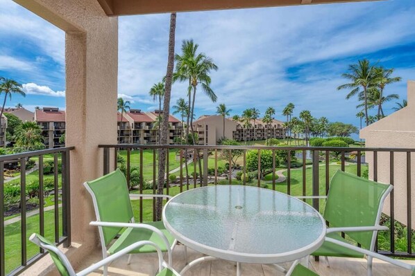 A balcony with a glass table and two chairs, overlooking a well-maintained lawn and palm trees, with buildings in the background. - Kamaole Sands (Kihei)