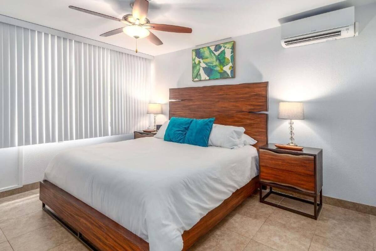 A modern bedroom with a wooden bed, white bedding, a ceiling fan, and an air conditioning unit.
