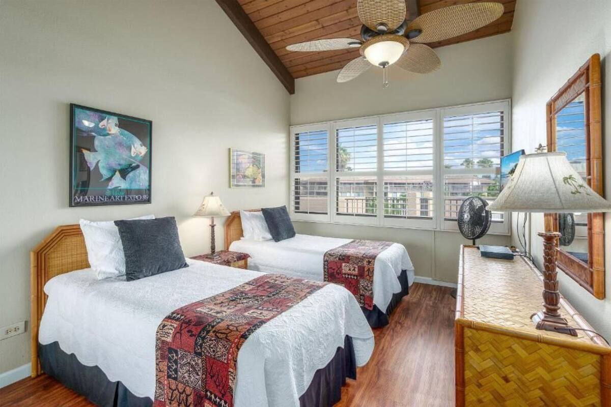 A hotel room with two beds, a ceiling fan, a large window with blinds, a wooden floor, and a framed picture on the wall.