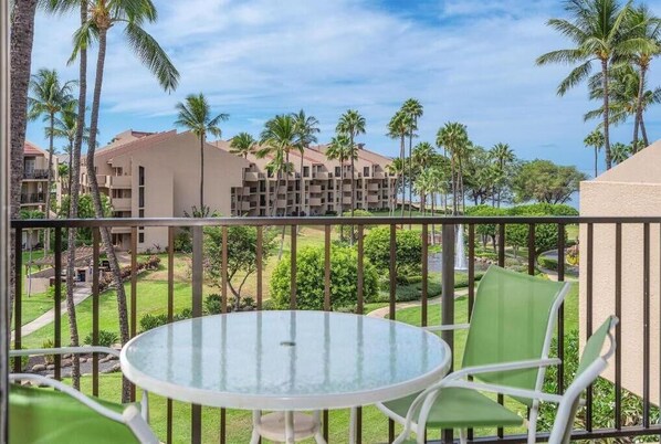 A balcony with a glass table and chairs, overlooking a resort with palm trees and a clear sky. - Kamaole Sands (Kihei)