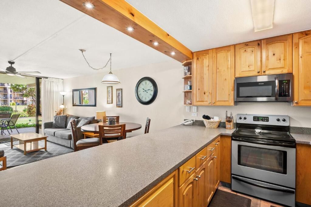 A modern kitchen with wooden cabinets, a microwave, and an oven. There is a dining area with a table and chairs, and a clock on the wall.