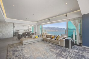 A modern living room with a large sectional sofa, a dining area, and a kitchen. There is a large window offering a view of the mountains.
