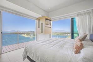 A bedroom with a large bed, a built-in TV, and a balcony with a view of the sea and a bridge.