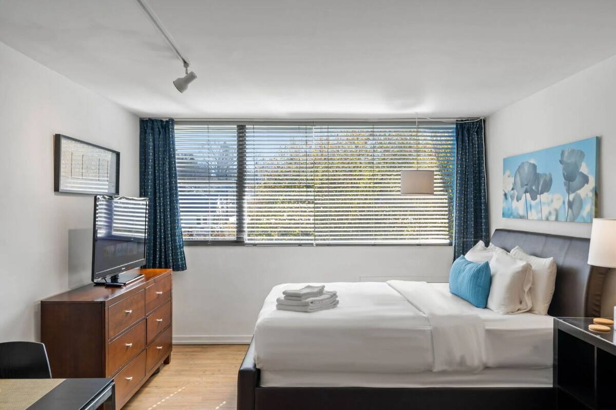 A hotel room with a bed, a desk, a dresser, a television, and a window with blinds.