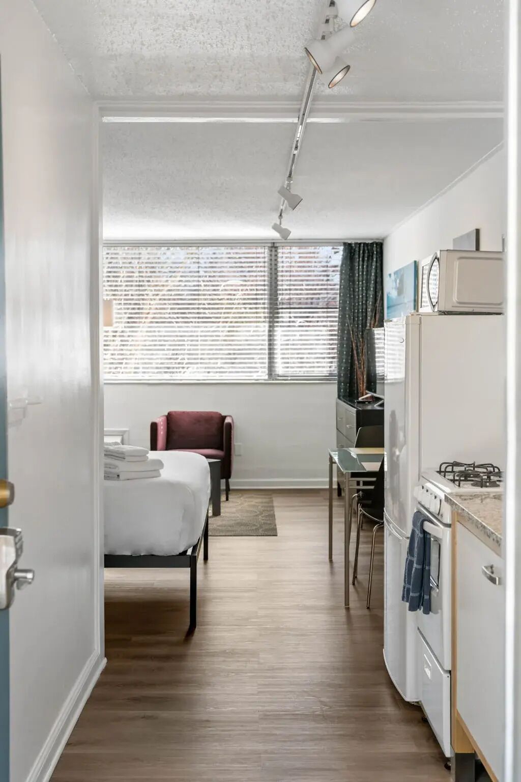 A compact hotel room with a bed, a small desk, a chair, a kitchenette with a stove, and a window with blinds.