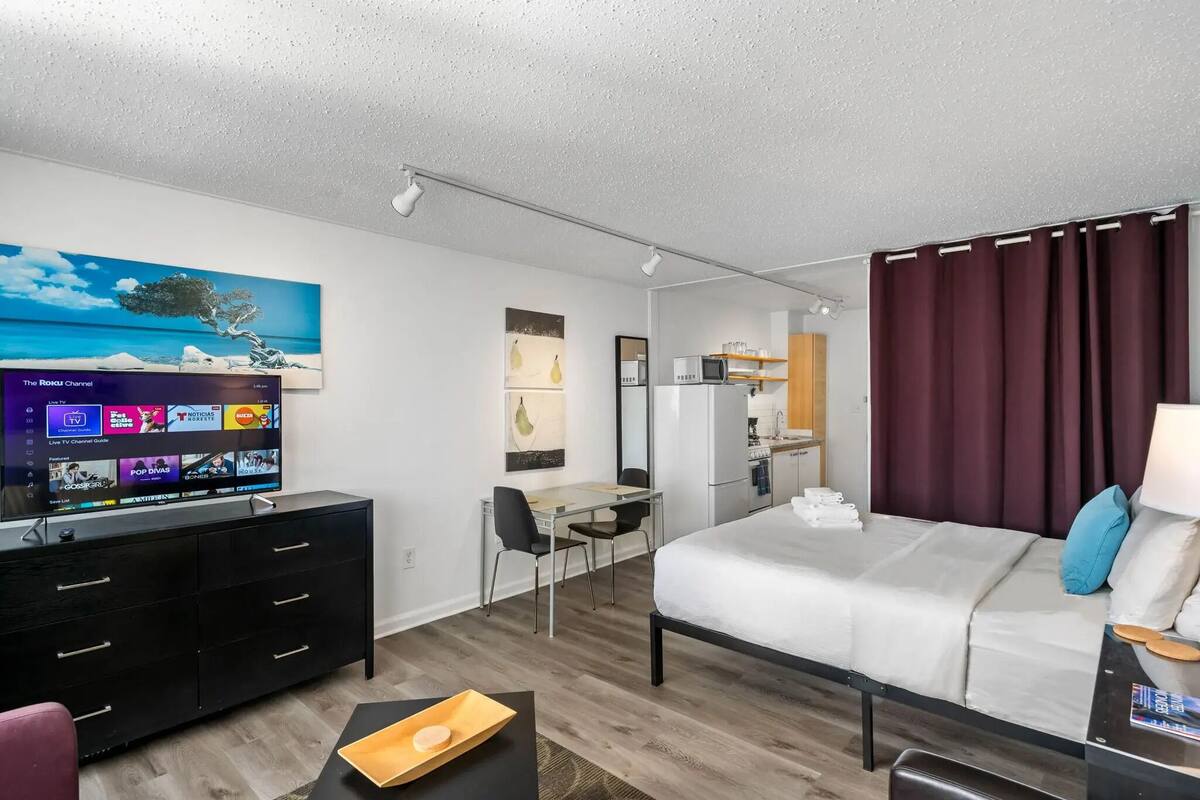 A modern hotel room with a bed, a desk, a TV, and a kitchen area.