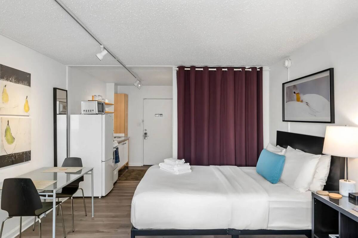 A hotel room with a bed, a desk, a chair, a microwave, a refrigerator, and a door leading to a kitchenette.