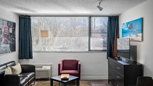 Habitación de hotel con un ventanal grande, un sofá, una silla, una mesita, un aparador y un cuadro en la pared.
