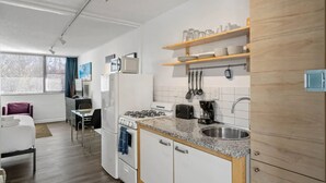 Cocina compacta con estufa a gas, microondas y cafetera. Hay estantes de madera sobre el lavamanos y una ventana con persianas al fondo.