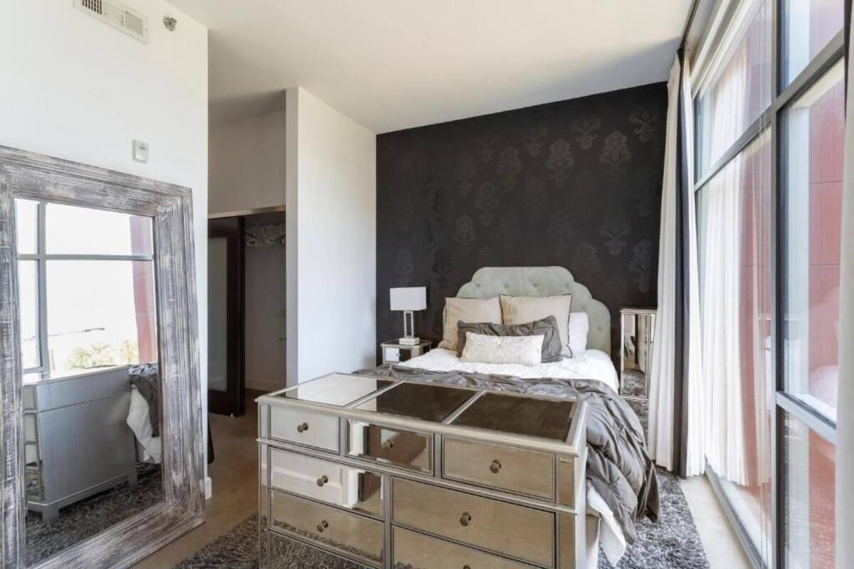 A bedroom with a large mirror, a bed with multiple pillows, a nightstand, and a dresser.