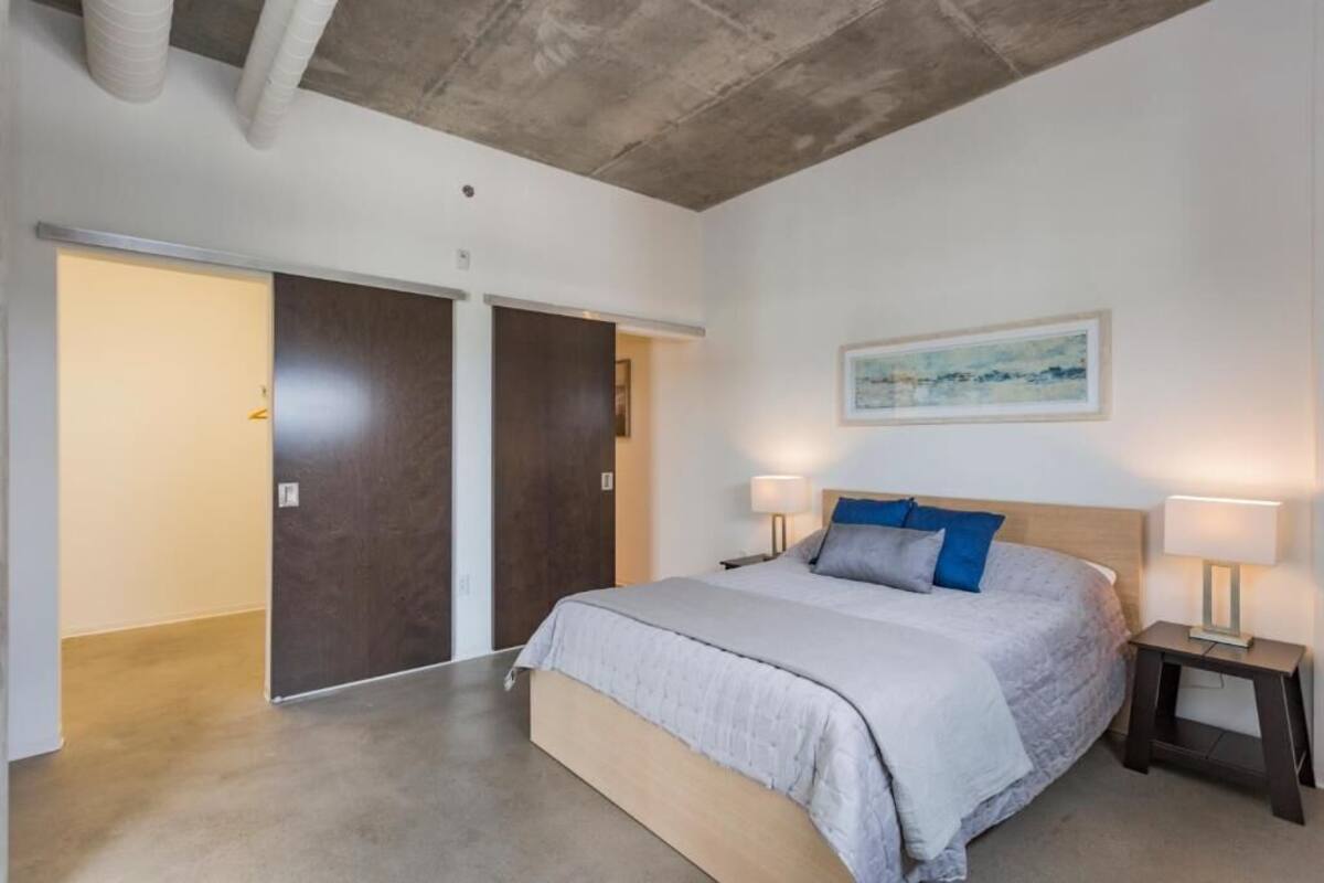 A bedroom with a large bed, two bedside tables with lamps, a wooden headboard, a painting on the wall, and a sliding door.