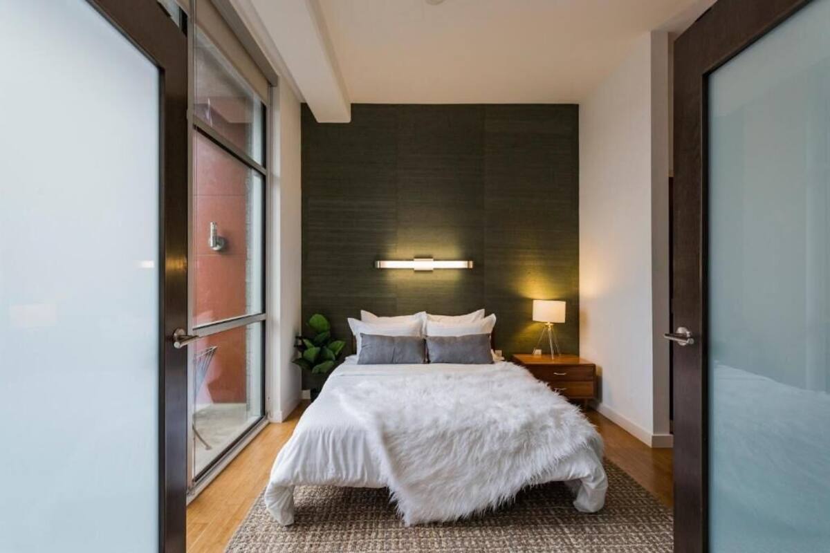 A modern bedroom with a large bed, a bedside table with a lamp, and a dark wall behind the bed.