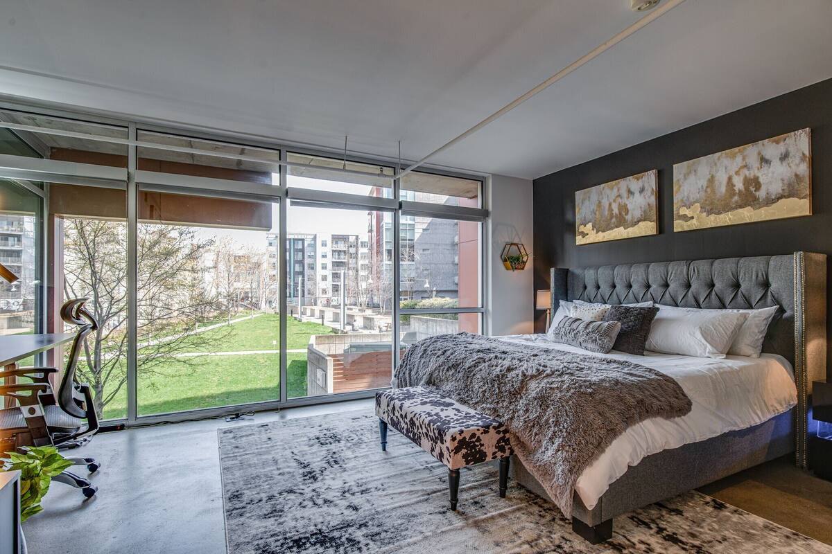 A modern bedroom with a large window, a tufted headboard, and a view of a cityscape.
