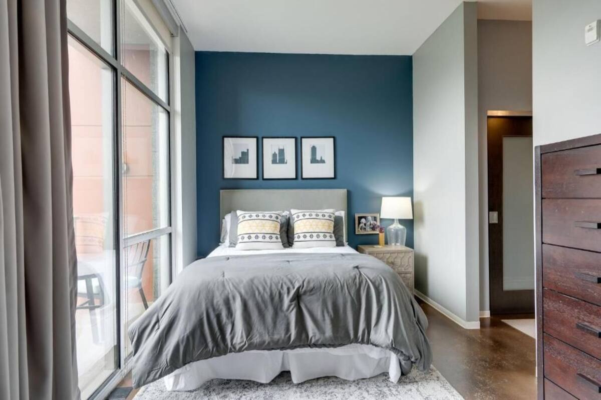 A bedroom with a large window, a bed with grey bedding, a nightstand with a lamp, and three framed pictures on the wall.