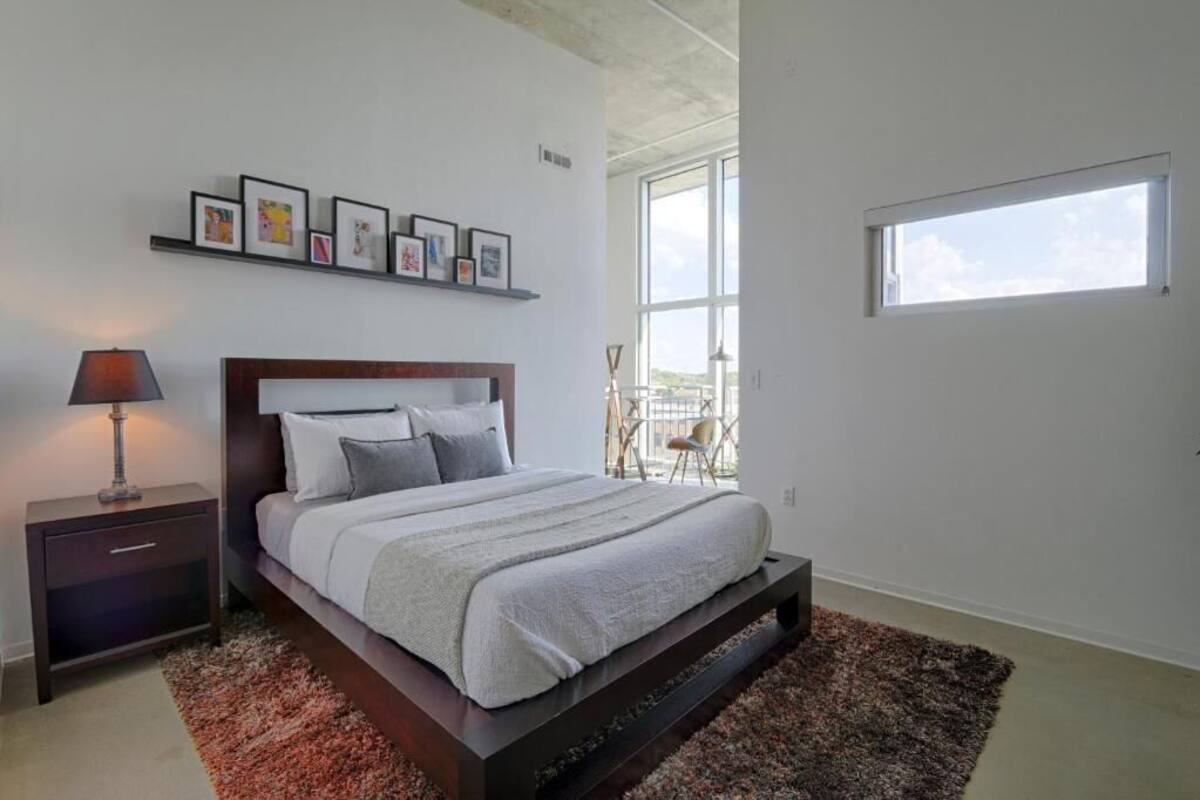 A modern bedroom with a large bed, a nightstand, a lamp, a framed picture collection, and a view of the outdoors through a window.